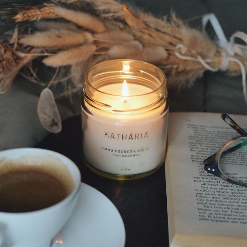 Foto af et tændt duftlys fra Kathária, kaffekop med kaffe, en åben bog med briller på.
